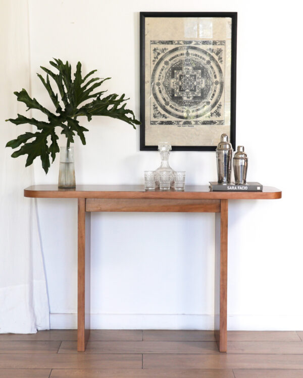 Consola London de lenga estilo elegante, apoyada contra pared blanca con cuadro enmarcado y decoración con jarrón de hojas verdes y copas.