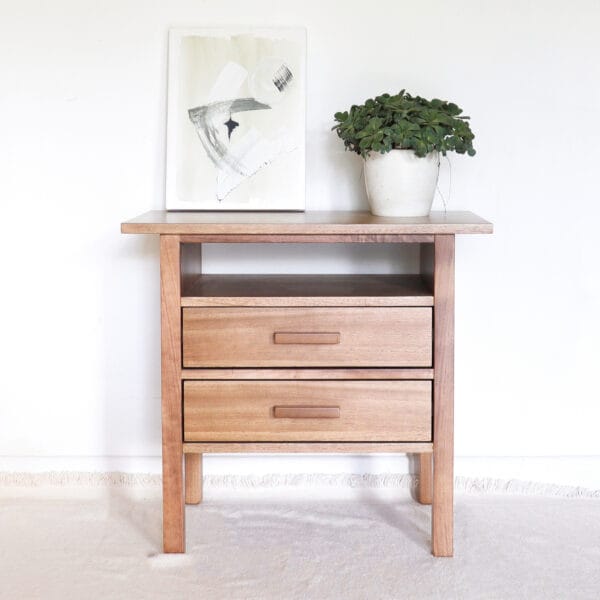 Mesa de luz Estocolmo de madera kiri, con diseño limpio y funcional, dos cajones amplios y un estante abierto, decorada con planta en maceta y cuadro minimalista sobre la tapa.