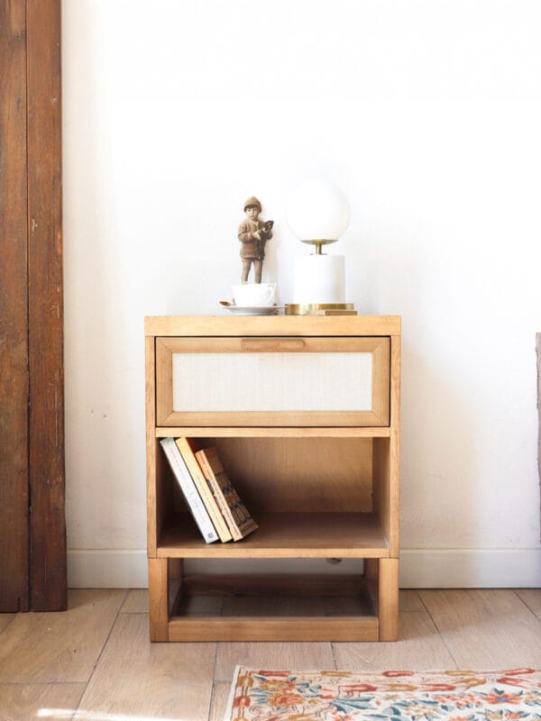 Mesa de luz Berna de madera clara con un cajón frontal de tela y dos estantes abiertos, decorada con una lámpara de diseño y libros.