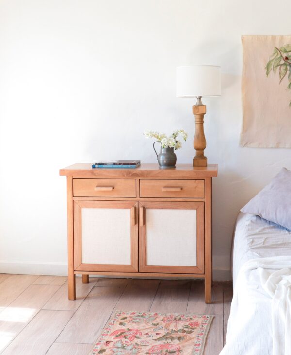 Cómoda Berna XS de madera kiri y rafia estilo cálido con dos cajones y puertas, vista en dormitorio con lámpara y flores decorativas.