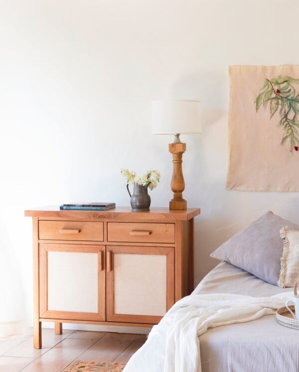 Cómoda Berna XS de madera kiri y rafia estilo cálido, mueble auxiliar con diseño nórdico cálido junto a cama matrimonial.