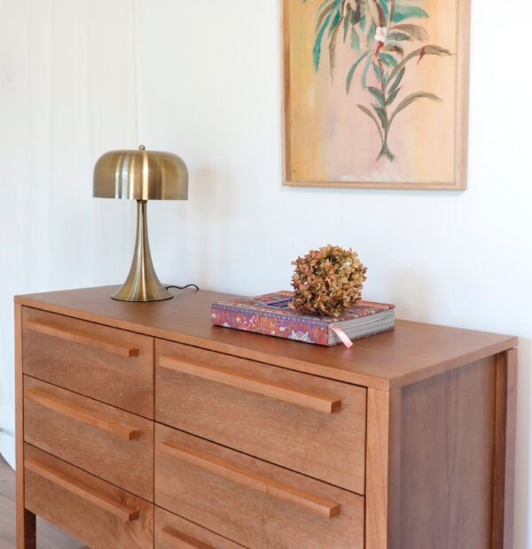 Cómoda Barcelona de madera kiri estilo moderno con lámpara dorada y cuadro decorativo en ambiente minimalista.