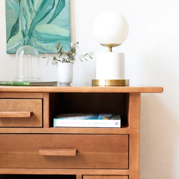 Detalle superior de la mesa de luz Bradbury de madera de lenga, estilo funcional, con lámpara de base dorada y esfera blanca, acompañada de un libro y un adorno sobre su superficie.