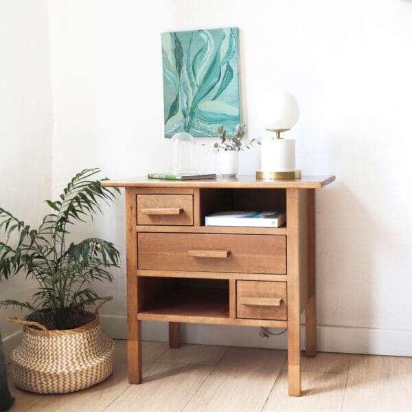 Vista completa de la mesa de luz Bradbury de madera de lenga, estilo funcional, con varios cajones de distintos tamaños y un estante abierto, acompañada de una planta decorativa al costado.