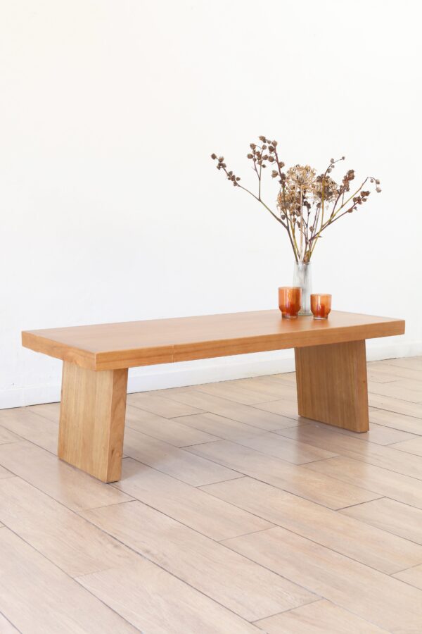 Mesa de living Serbia de madera kiri, diseño robusto con patas rectangulares anchas, ambientada con jarrón transparente con flores secas y dos vasos color ámbar sobre piso de madera clara.