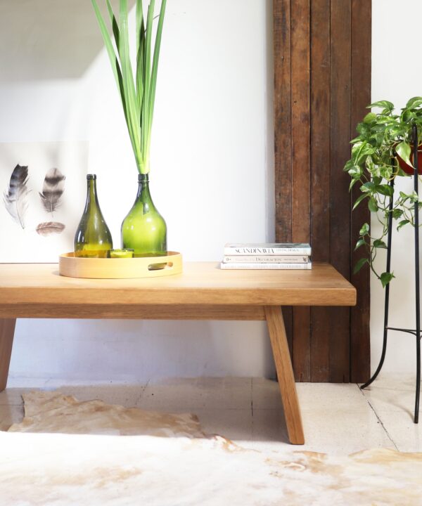 Mesa de living Serbia de madera kiri, estilo contemporáneo, con base inclinada y superficie rectangular, decorada con bandeja redonda de madera, botellas verdes y libros sobre un piso claro.
