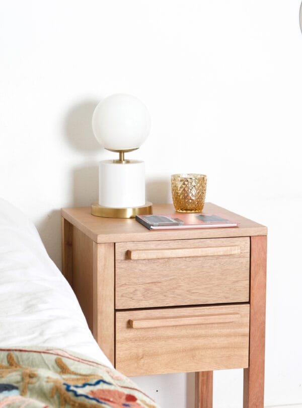 Mesa de luz Barcelona de madera kiri con dos cajones, diseño esbelto y funcional. Sobre la mesa hay una lámpara de base blanca y dorada con pantalla esférica, y un portavelas dorado decorativo.