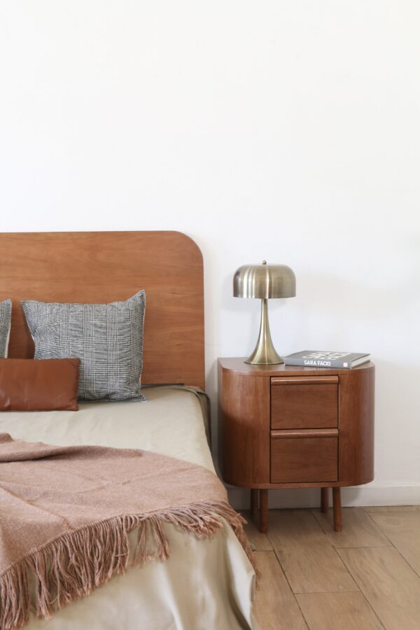 Mesa de luz London junto a cama matrimonial, fabricada en madera natural con líneas curvas y estilo contemporáneo.