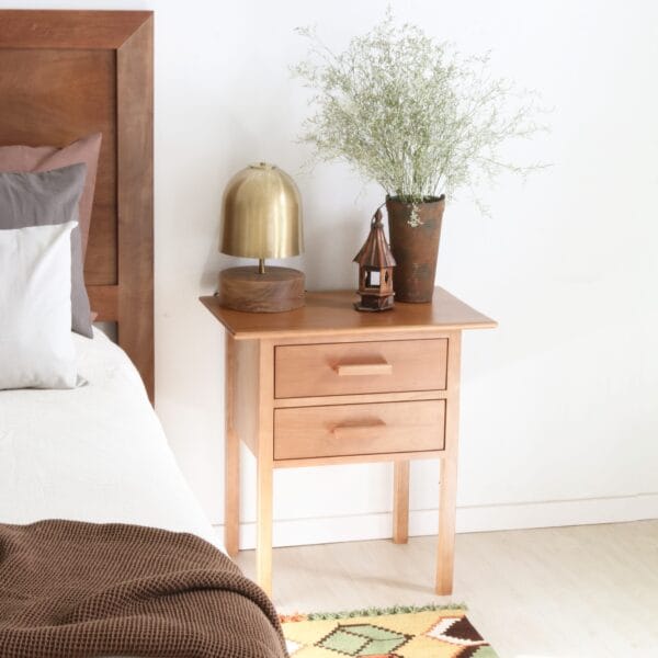 Mesa de luz Máximo de lenga con dos cajones, acompañada de lámpara dorada, adorno artesanal y florero alto, ambientada en dormitorio con estilo cálido