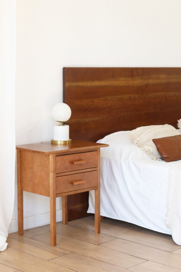 Mesa de luz Máximo de madera lenga, diseño clásico con dos cajones y tiradores rectos, ubicada junto a la cama con lámpara moderna de base dorada y pantalla esférica blanca