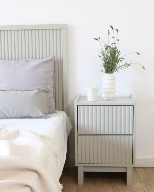 Mesa de luz Venecia en MDF gris con diseño moderno y ranurado vertical, decorada con jarrón blanco y lavanda.