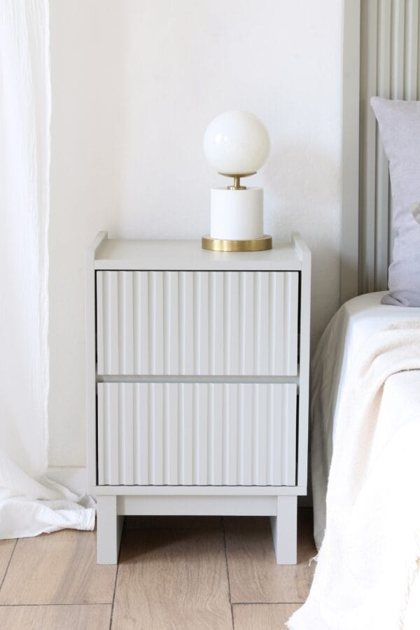 Mesa de luz Venecia moderna de MDF gris, con dos cajones de frente ranurado y lámpara de mesa dorada y blanca.