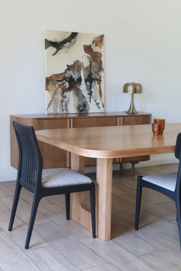 Detalle de la mesa de comedor London en madera kiri con sillas negras tapizadas en tono claro y aparador de fondo