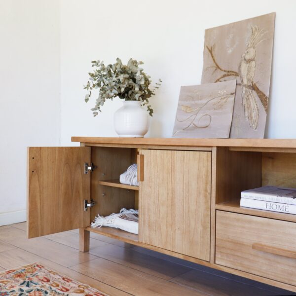 Detalle del aparador Oslo de madera kiri estilo escandinavo, con puerta abierta mostrando estantes internos y decoración con cuadros y jarrón de cerámica.