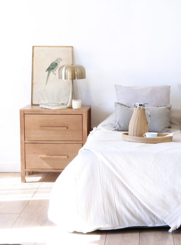 Mesa de luz Provance de madera kiri con dos cajones, ubicada junto a la cama, decorada con lámpara dorada, cuadro de ave y detalles de desayuno sobre la cama.