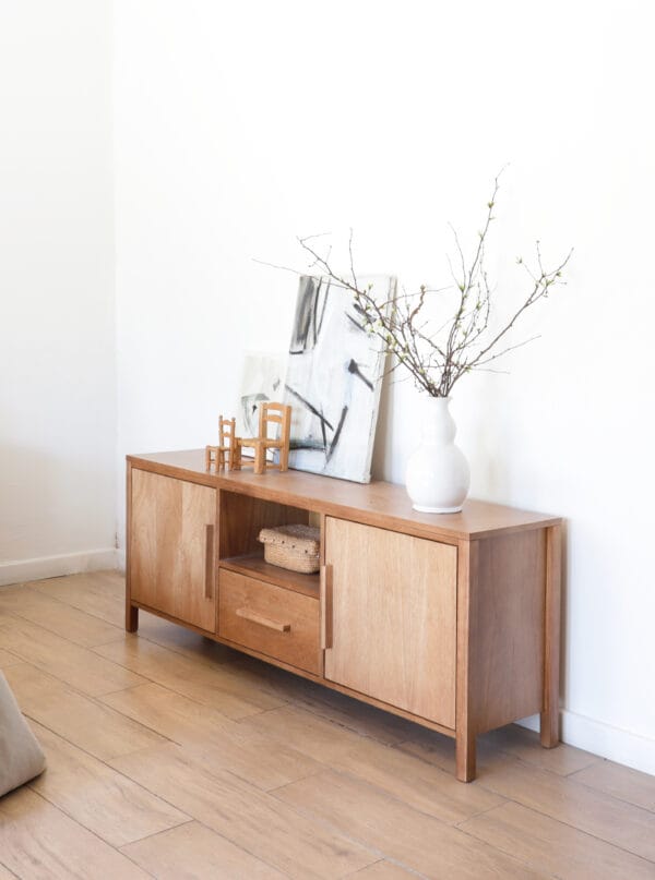 "Aparador Provance chico en madera kiri con diseño limpio y práctico, decorado con jarrón blanco y cuadros modernos sobre su superficie.