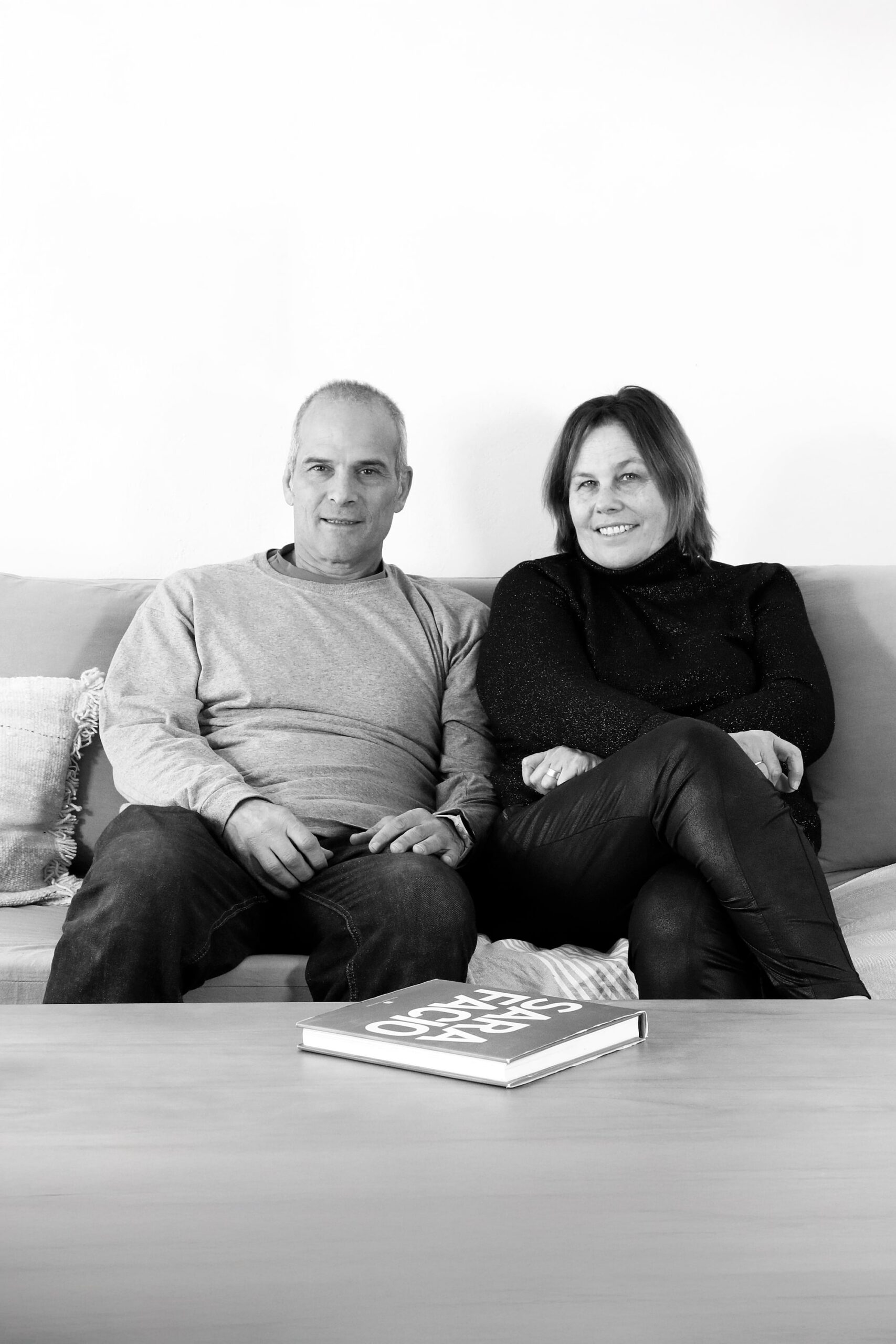 Una pareja, un hombre y una mujer, están sentados en un sofá y miran a la cámara. Un libro de tapa dura está sobre la mesa frente a ellos. La imagen está en blanco y negro.