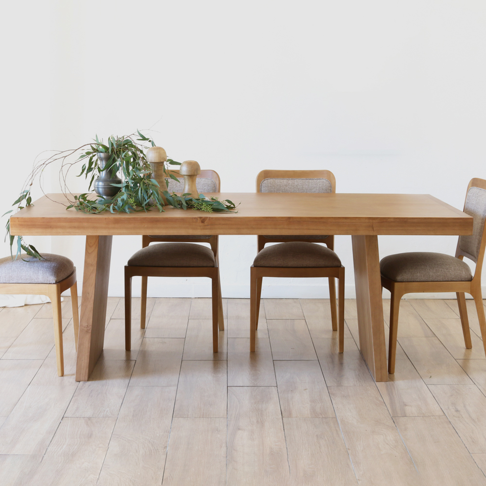 Mesa de madera larga con sillas de madera y tapicería, en un comedor minimalista con suelo de madera. Un jarrón con ramas de eucalipto y adornos de madera adornan el centro de la mesa.