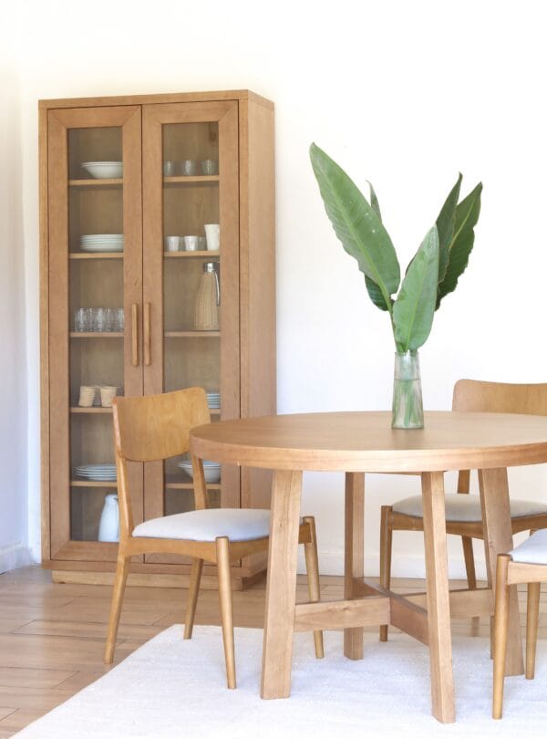 Mesa de comedor Provance redonda en madera kiri estilo campiña francesa con sillas tapizadas en tono claro y aparador de fondo con vajilla